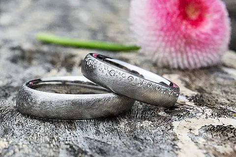 Eheringe Platin oder Weißgold mit einer eismatten Oberfläche und verschieden großen Steinen (Brillanten) im Damenring die wie ein Zuckerrand willkürlich auf einer Seite verteilt sind