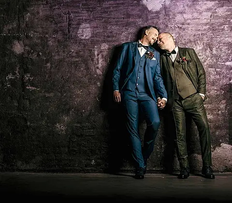 Zwei Männer in Hochzeitsanzügen lehnen an einer alten Mauer verliebt aneinander
